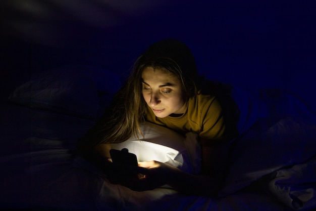 Una chica en la cama con un teléfono en una cama blanca en una habitación oscura