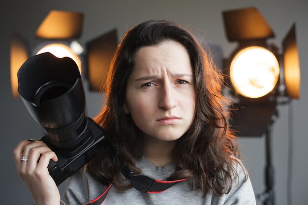 Chica con una cámara en el estudio sobre la iluminación de fondo Ella frunce el ceño y mira hacia adelante con dudas