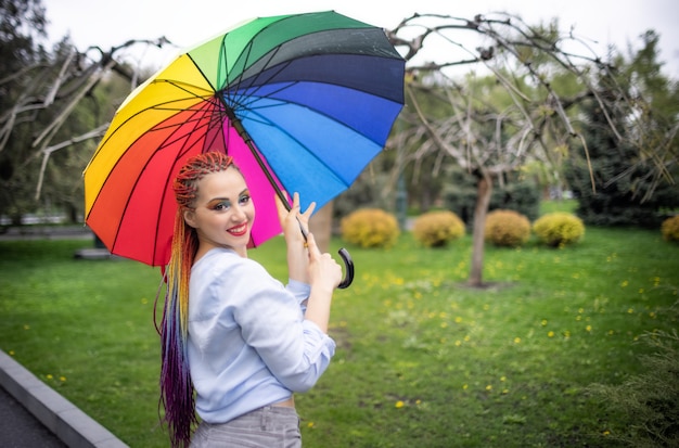 Chica con una camisa azulada con maquillaje brillante y largas trenzas de colores. Sonriendo y sosteniendo un paraguas con los colores del arco iris en el fondo de un parque florecido disfrutando de la próxima primavera.