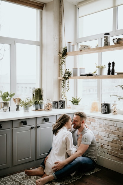 Chica con una camisa blanca y un chico con una camiseta gris en la cocina. Besos y abrazos.