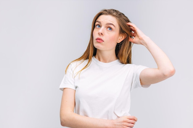 Chica en camisa blanca duda rascándose la cabeza sobre fondo gris aislado