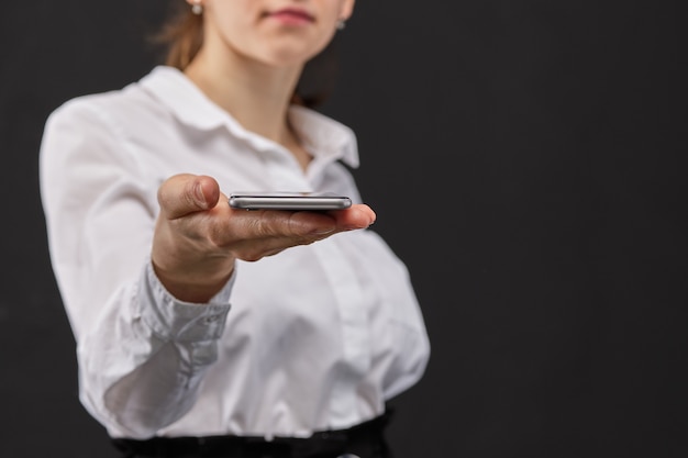 Chica con una camisa blanca extiende su mano con un teléfono inteligente sobre un fondo negro.