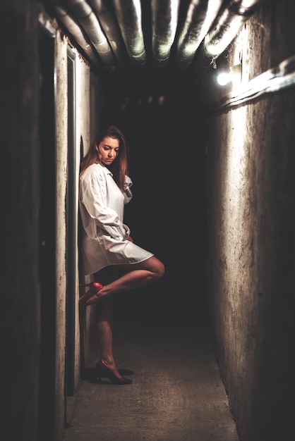 Chica con camisa blanca larga y tacones altos posando en un pasillo oscuro