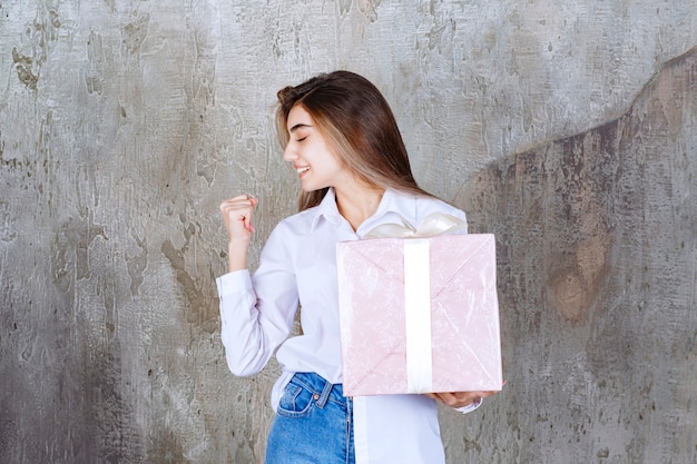 Chica con camisa blanca sosteniendo una caja de regalo rosa envuelta con cinta blanca y mostrando un signo de mano positivo.