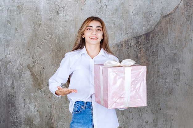 Chica con camisa blanca sosteniendo una caja de regalo rosa envuelta con una cinta blanca, notando a su pareja y pidiéndole que venga a recibirla.