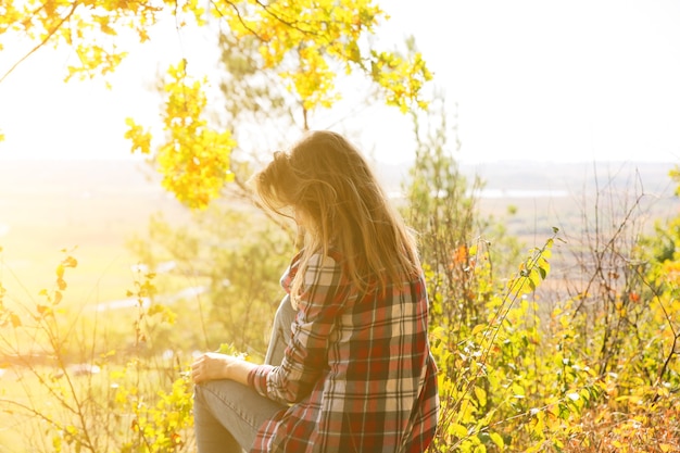 Chica de la camisa a cuadros está sentada en el bosque de otoño. Concepto estacional. Ropa con estilo hipster al aire libre. Filosofía de la naturaleza alrededor. Mujer hermosa cerca de los árboles y las hojas amarillas.