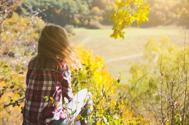 Chica de la camisa a cuadros está sentada en el bosque de otoño. Concepto estacional. Ropa con estilo hipster al aire libre. Filosofía de la naturaleza alrededor. Mujer hermosa cerca de los árboles y las hojas amarillas.