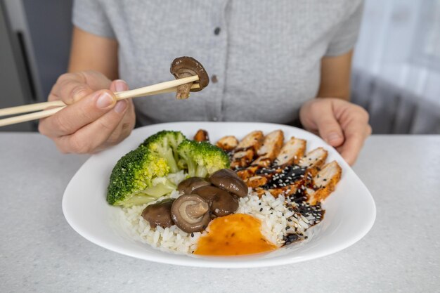 Una chica con una camisa gris en la cocina de casa sostiene un plato de arroz dorado con champiñones shiitake, pollo con salsa unagi y brócoli en sus manos Concepto de comida asiática