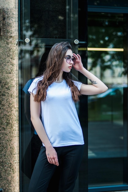 Una chica con una camiseta blanca en blanco y jeans se para cerca de una maqueta de pared de vidrio para la imprenta de camisetas