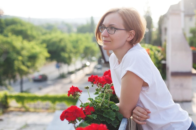 Una chica con una camiseta blanca y gafas se para en el balcón y disfruta de la belleza y el cuidado. luz solar brillante.