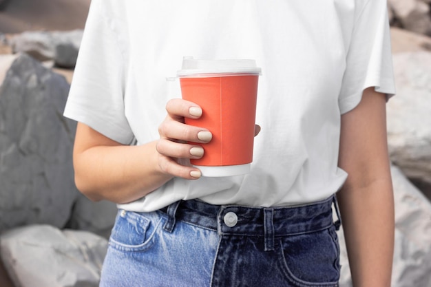 Una chica con una camiseta blanca y jeans azules sostiene una taza roja para tomar café contra el fondo de una playa de arena y rocas grises