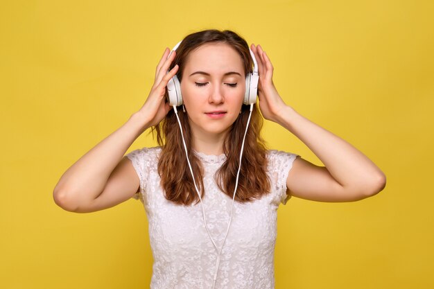 Una chica con una camiseta blanca y jeans marrones sobre un fondo amarillo se relaja y escucha atentamente y disfruta de la música en los auriculares blancos cerrando los ojos.