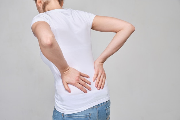 Una chica con una camiseta blanca se masajea la zona lumbar por un dolor intenso. Ciática, sciosis.