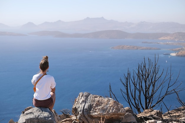 Chica con una camiseta blanca sentada con la espalda en una montaña de roca de piedra con vistas al mar y la montaña