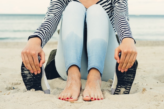 Chica en camiseta a rayas y jeans se sienta descalza en la playa al lado de los zapatos