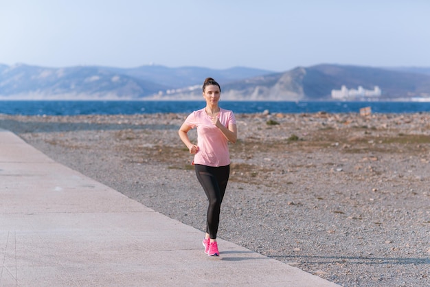 una chica con una camiseta rosa y polainas negras hace deporte en la orilla del mar