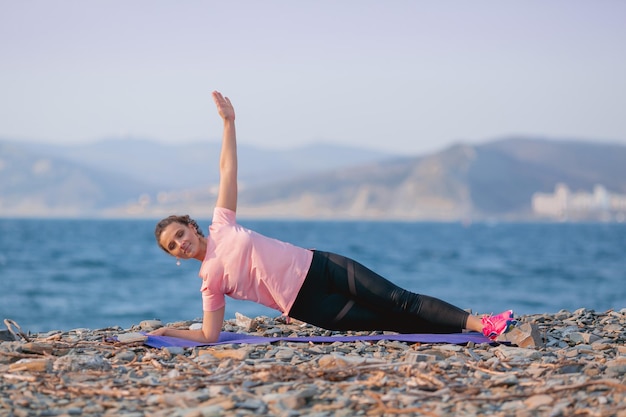 una chica con una camiseta rosa y polainas negras hace deporte en la orilla del mar