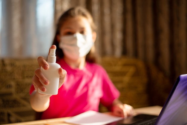 Chica en camiseta rosa rociando desinfectante para manos mientras trabajaba en una computadora portátil, estudiando en casa durante la cuarentena de coronavirus