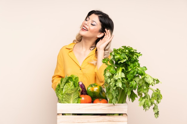 Chica campesina adolescente con verduras recién cortadas en una caja en la pared de color beige escuchando algo