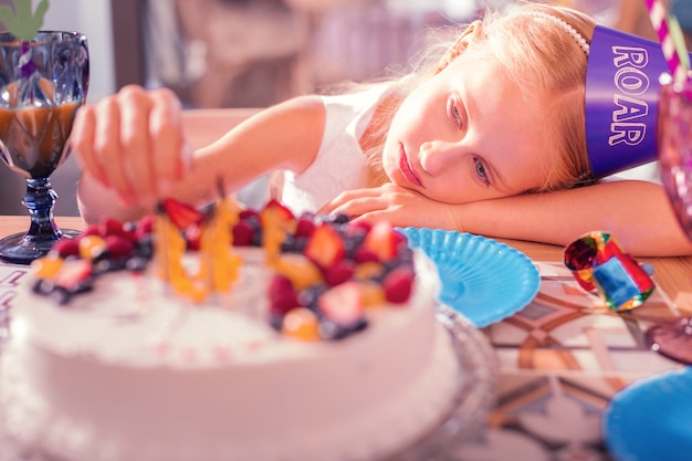 Chica cansada sentada en la mesa y mirando su pastel de cumpleaños