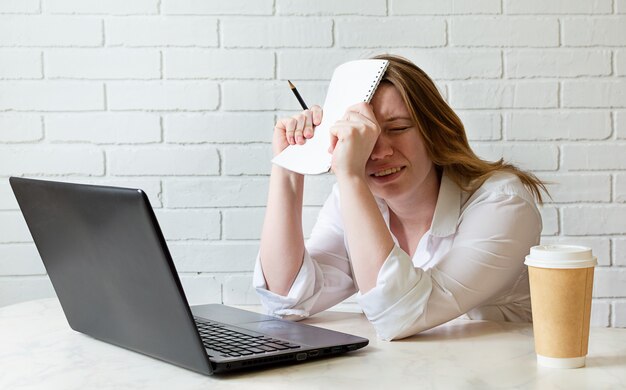 Chica cansada sentada en la mesa de trabajo en casa estudiando en el portátil con café