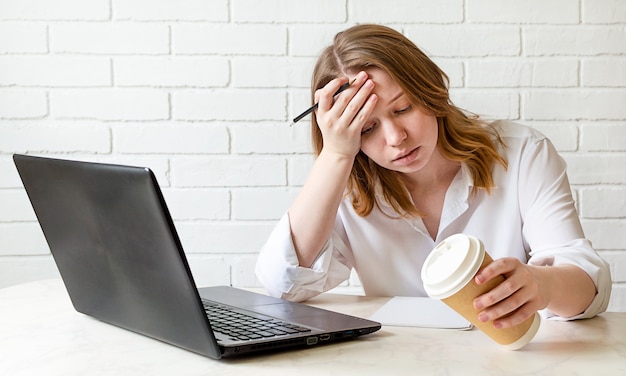 Chica cansada sostiene su cabeza con la mano y sentado en la mesa de trabajo en casa estudiando en la computadora portátil con café