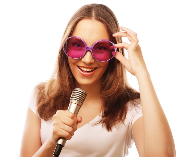 Chica cantando feliz. Mujer de belleza con camiseta blanca y gafas de sol grandes con micrófono sobre fondo blanco. estilista inconformista