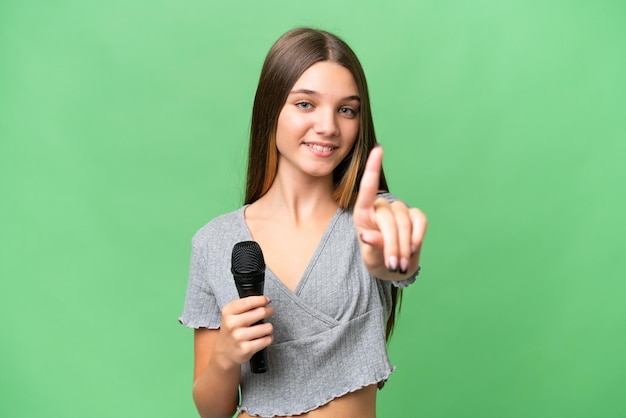 Foto chica cantante adolescente recogiendo un micrófono sobre un fondo aislado mostrando y levantando un dedo