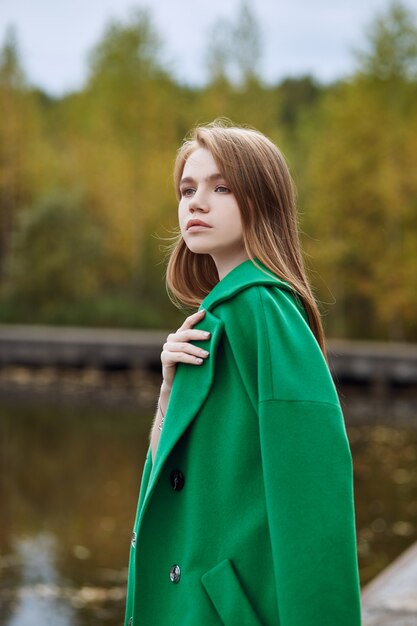 Chica en una capa verde camina a lo largo del terraplén del lago en un día nublado de otoño.