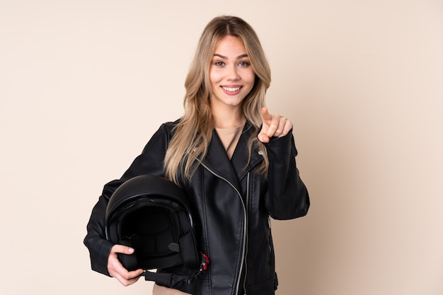 Chica con un casco de moto en pared beige sorprendido y señalando