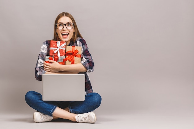 Una chica casual emocionada sosteniendo la computadora portátil mientras está sentado en el suelo con una pila de cajas de regalo