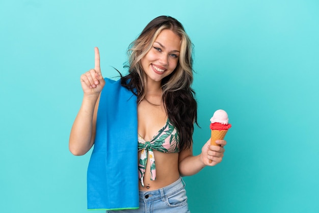 Chica caucásica adolescente sosteniendo helado y toalla aislada de fondo azul mostrando y levantando un dedo en señal de lo mejor