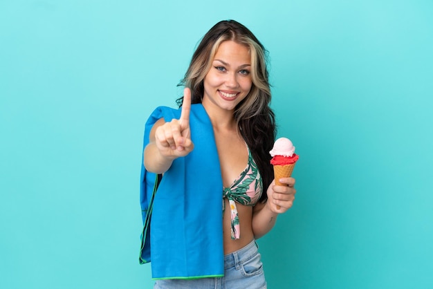 Chica caucásica adolescente sosteniendo helado y toalla aislada de fondo azul mostrando y levantando un dedo