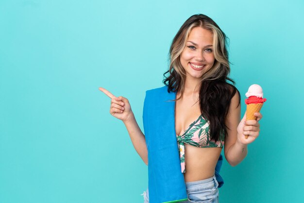 Chica caucásica adolescente sosteniendo helado y toalla aislado sobre fondo azul apuntando con el dedo al lado