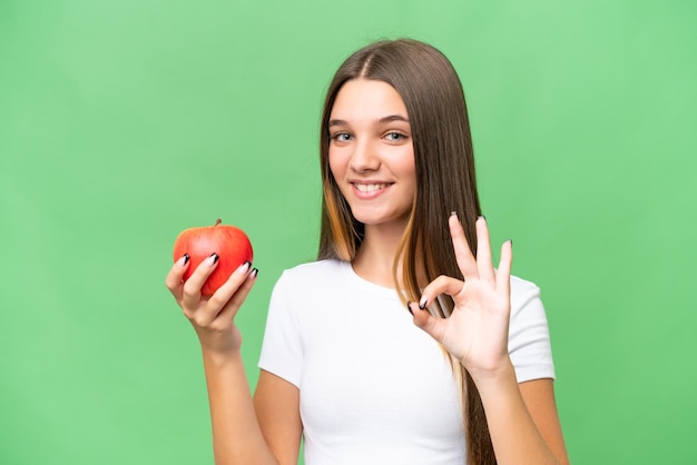 Chica caucásica adolescente sosteniendo una manzana sobre un fondo aislado que muestra el signo de ok con los dedos