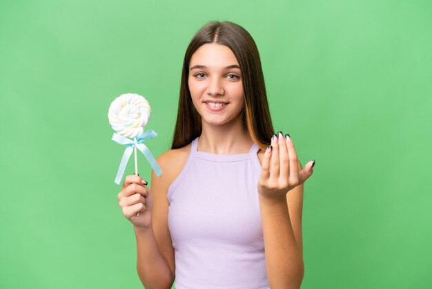 Chica caucásica adolescente sosteniendo una piruleta sobre un fondo aislado invitando a venir con la mano feliz de que hayas venido