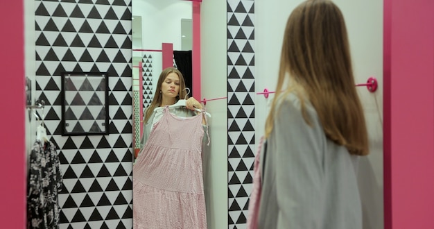 Foto chica caucásica bastante elegante probándose un bonito vestido en el espejo en el vestidor en la tienda de ropa. hermosa joven eligiendo ropa en elegante llaga de centro comercial.