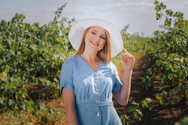 Foto chica caucásica feliz con vestido azul y sombrero sonriendo en un viñedo