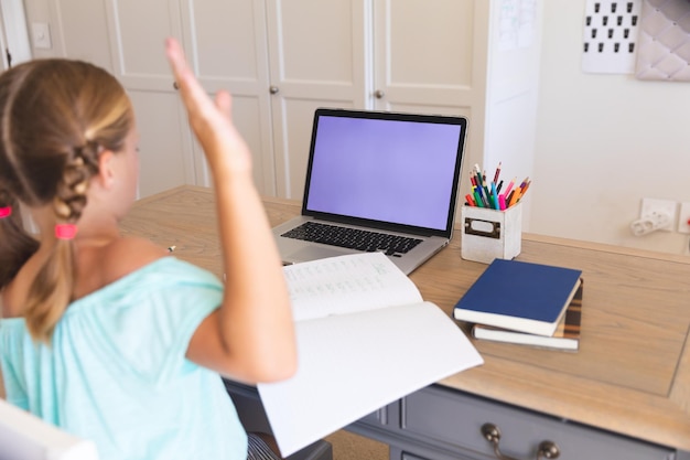 Chica caucásica haciendo videollamadas durante la clase en una laptop con espacio para copiar y levantando la mano en casa