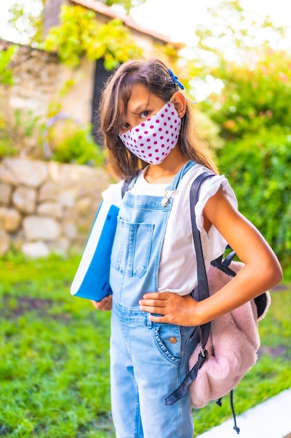 Chica caucásica con mascarilla lista para volver a la escuela. Nueva normalidad, distancia social, pandemia de coronavirus, covid-19. Chaqueta, mochila y bloque azul para notas en la mano