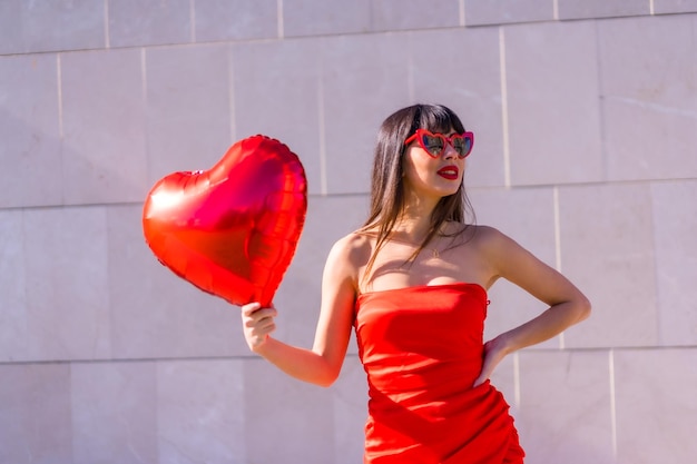 Chica caucásica morena con un globo de corazón en gafas de sol del día de San Valentín y vestido rojo Estilo de vida de personas enamoradas sobre un fondo gris