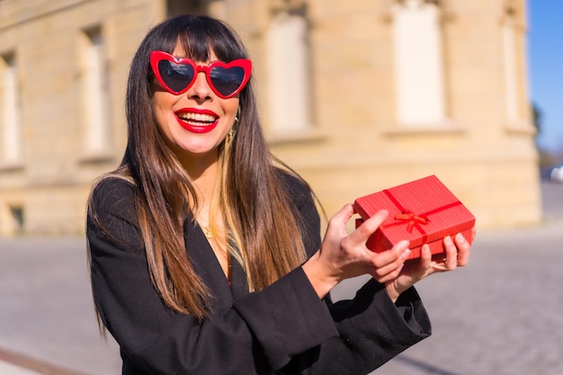 Chica caucásica morena con un regalo en el día de San Valentín gafas de sol de corazones rojos muy feliz con la sorpresa