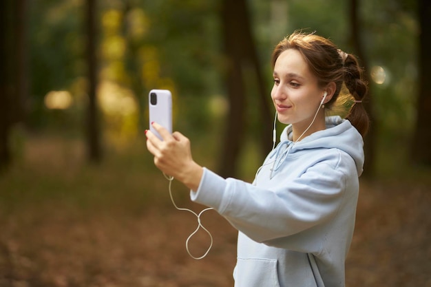 Una chica caucásica en el parque hablando en una videollamada a través de auriculares y un teléfono móvil