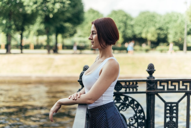 Una chica caucásica con un peinado bob camina en un parque de la ciudad sobre el puente y mira hacia un lado.