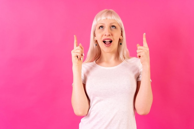 Chica caucásica rubia sobre fondo de estudio rosa sonriendo apuntando hacia el espacio de copia