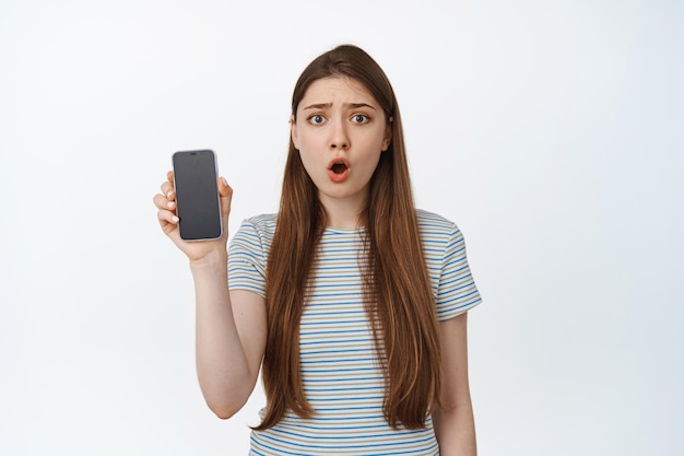 Chica caucásica sorprendida muestra la pantalla del teléfono inteligente con cara preocupada, reacción a algo molesto en el teléfono móvil, de pie contra el fondo blanco