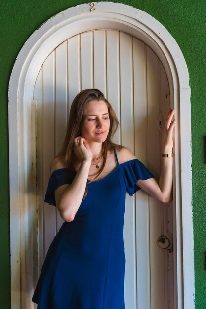 Chica caucásica con un vestido azul junto a una puerta blanca de una foto vertical de una casa verde