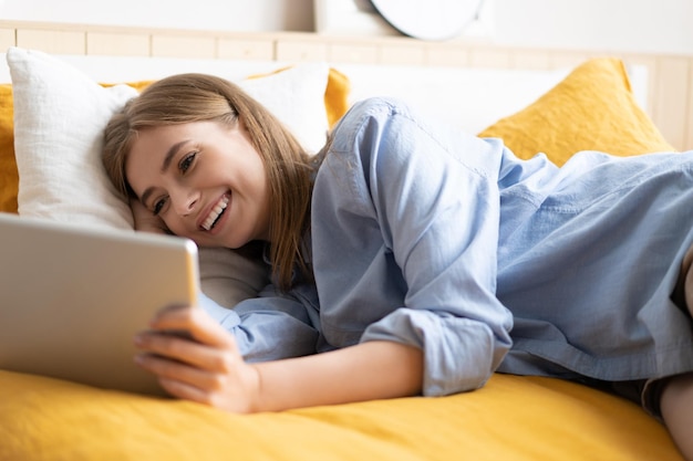 Chica caucásica con videochat con amigos usando la cámara de la tableta mientras está acostada en el concepto de comunicación de tecnología de cama