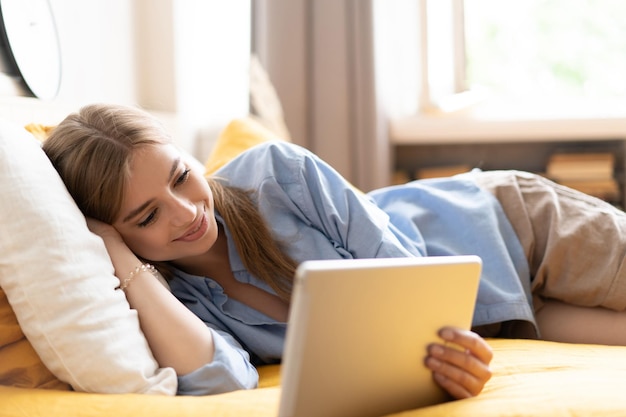 Chica caucásica con videochat con amigos usando la cámara de la tableta mientras está acostada en el concepto de comunicación de tecnología de cama