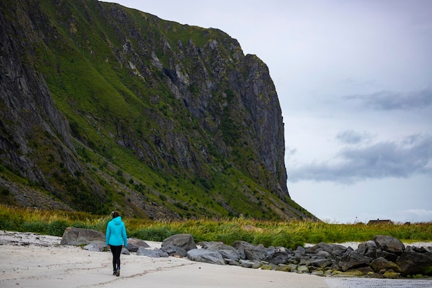 chica con chaqueta azul camina por la playa con enormes rocas y montañas al fondo, Lofoten